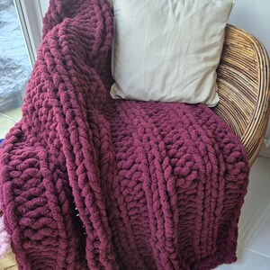 May include: A chunky knit throw blanket in a deep burgundy color draped over a wicker chair with a cream colored pillow.