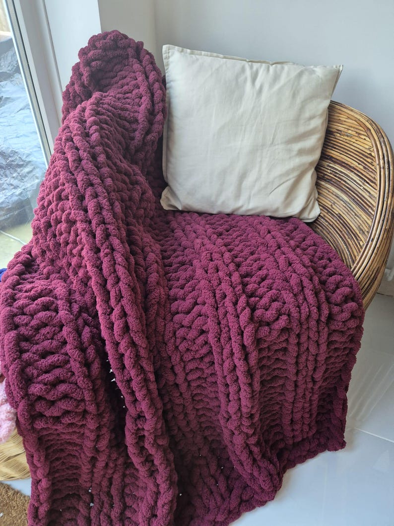 May include: A chunky knit throw blanket in a deep burgundy color draped over a wicker chair with a cream colored pillow.
