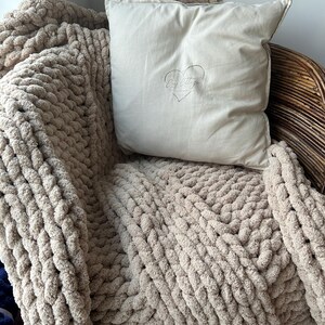 May include: A beige chunky knit throw blanket draped over a wicker chair with a cream colored pillow featuring the text "By Little" and a heart design.