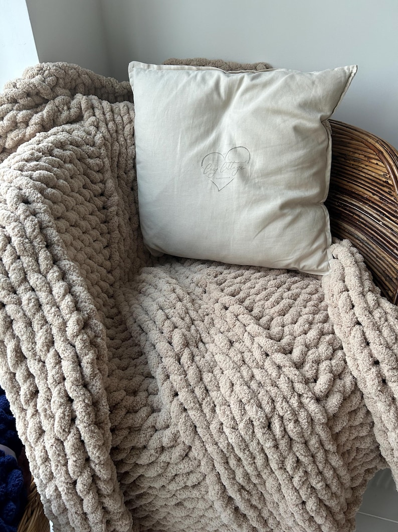 May include: A beige chunky knit throw blanket draped over a wicker chair with a cream colored pillow featuring the text "By Little" and a heart design.