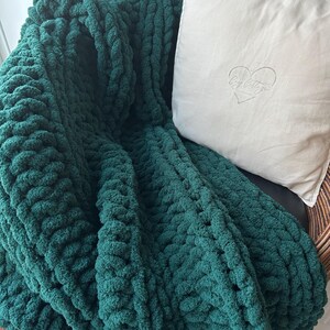 May include: A deep green chunky knit throw blanket draped over a chair with a white pillow featuring the text "Cozy Cottage" in a heart shape.