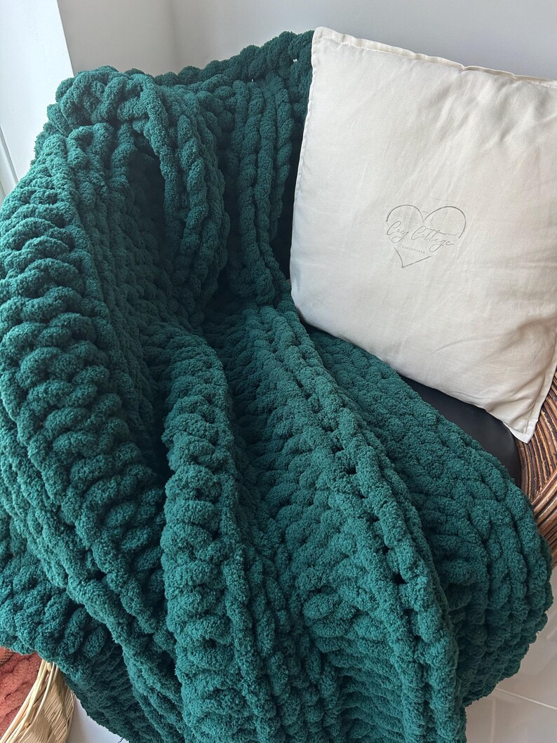 May include: A deep green chunky knit throw blanket draped over a chair with a white pillow featuring the text "Cozy Cottage" in a heart shape.