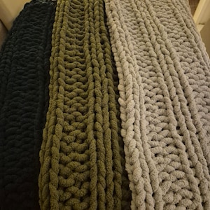 May include: Three chunky knit blankets in black, olive green, and light gray. The blankets are made with thick yarn and have a textured, cable knit pattern.