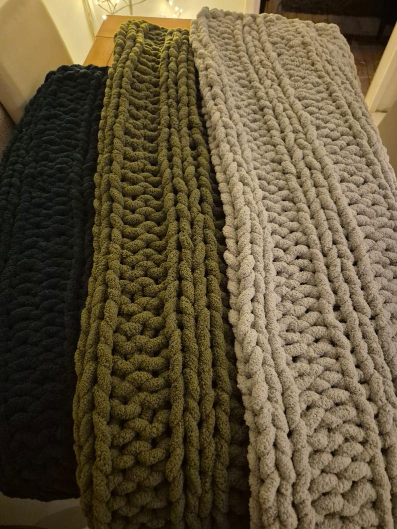 May include: Three chunky knit blankets in black, olive green, and light gray. The blankets are made with thick yarn and have a textured, cable knit pattern.