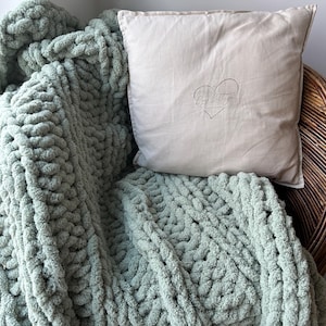 May include: A sage green chunky knit throw blanket draped over a wicker chair with a beige pillow featuring the text "My Little Cottage" and a heart design.