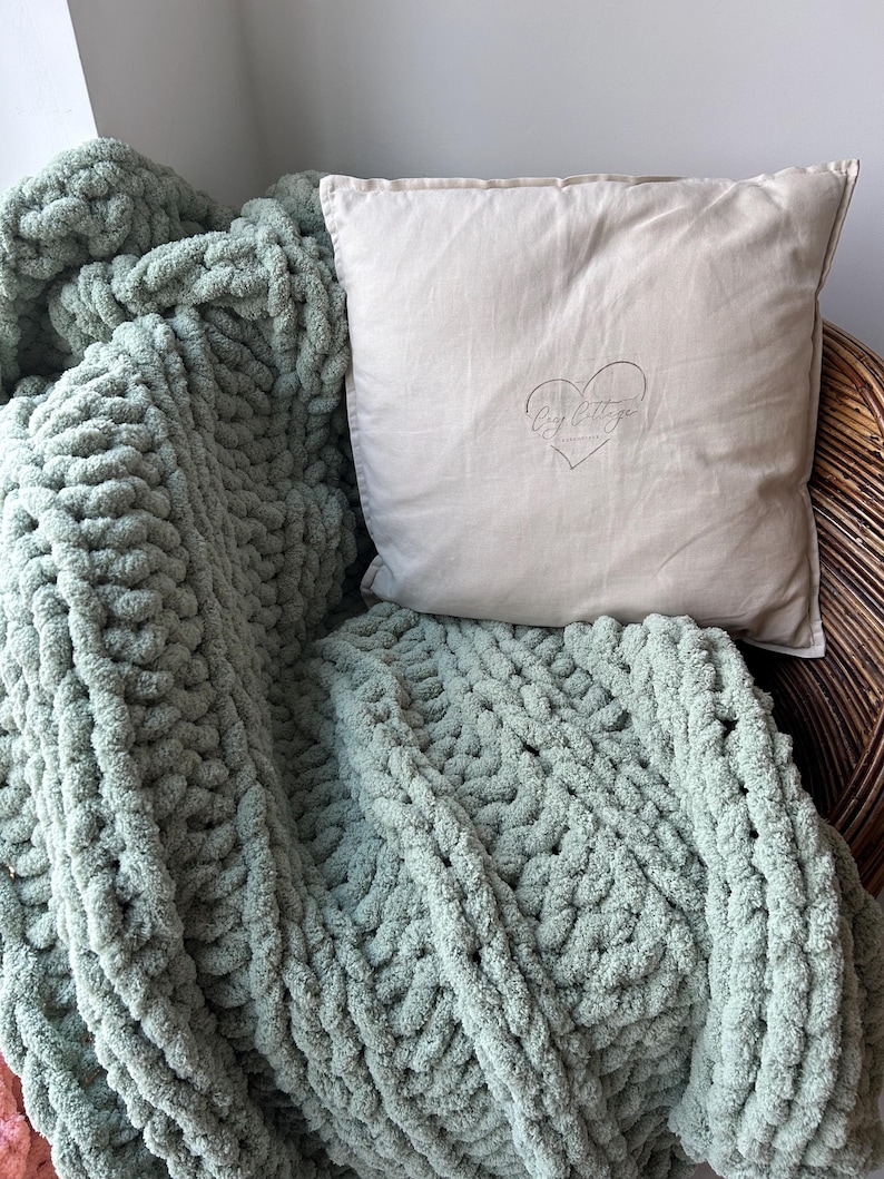 May include: A sage green chunky knit throw blanket draped over a wicker chair with a beige pillow featuring the text "My Little Cottage" and a heart design.