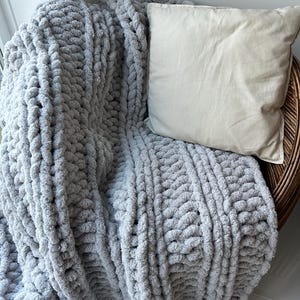 May include: A gray chunky knit throw blanket draped over a wicker chair with a beige pillow. The blanket has a textured, cable knit pattern.