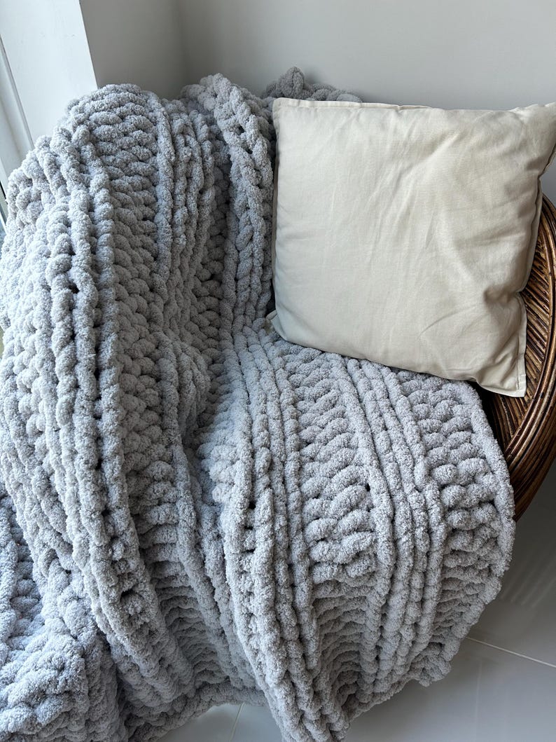 May include: A gray chunky knit throw blanket draped over a wicker chair with a beige pillow. The blanket has a textured, cable knit pattern.
