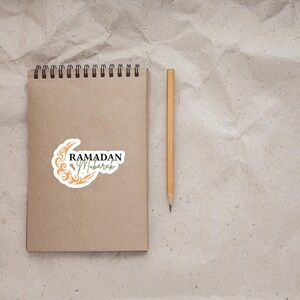 Pu&ograve; includere: Un quaderno marrone con una rilegatura a spirale e un adesivo che dice "Ramadan Mubarak" con un disegno di luna crescente e stelle. Una matita gialla &egrave; accanto al quaderno.