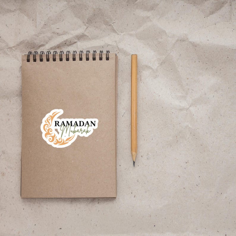 Pu&ograve; includere: Un quaderno marrone con una rilegatura a spirale e un adesivo che dice "Ramadan Mubarak" con un disegno di luna crescente e stelle. Una matita gialla &egrave; accanto al quaderno.