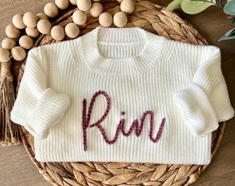 May include: White knitted sweater with a maroon embroidered name "Rin" on the front.
