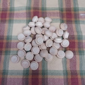 Puede incluir: Un conjunto de tapones de madera sin terminar, cada uno con una parte superior redondeada y una base ligeramente más ancha. Los tapones están esparcidos sobre un fondo de tela a cuadros en tonos verdes, rojos y beige. La madera es de color claro natural.