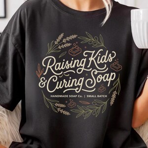 Peut inclure: T-shirt noir de couleur confort avec l'inscription "Raising Kids & Curing Soap" dans un motif circulaire. Le motif comprend des illustrations de savon et des éléments botaniques. Produit artisanal de petite série.