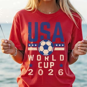 May include: Red t-shirt with the text "USA WORLD CUP 2026" in blue and white. The design includes a soccer ball and stars. The person is holding a sparkler and a small American flag.