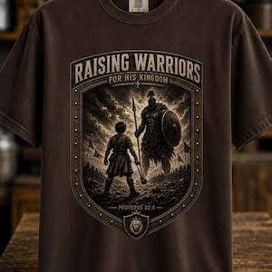May include: A dark brown short-sleeve t-shirt featuring a graphic print of a child standing before a large warrior figure. The text reads Raising Warriors For His Kingdom and Proverbs 22:6. The design is enclosed in a shield-shaped border.