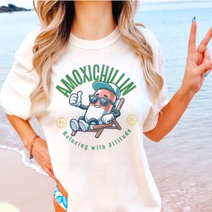 May include: A white t-shirt with a green and red cartoon pill wearing sunglasses and a hat, sitting in a chair. The text "Amoxichillin Relaxing with Altitude" is printed on the shirt.