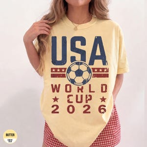 May include: Butter yellow t-shirt with a distressed USA World Cup 2026 graphic. The design features a soccer ball, stars, and the text in navy blue and red. The shirt is a comfort color.