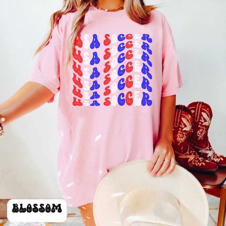 May include: A pink oversized t-shirt with a graphic print of "USA SOCCER" repeated in red, white, and blue. The graphic is in a retro font style.