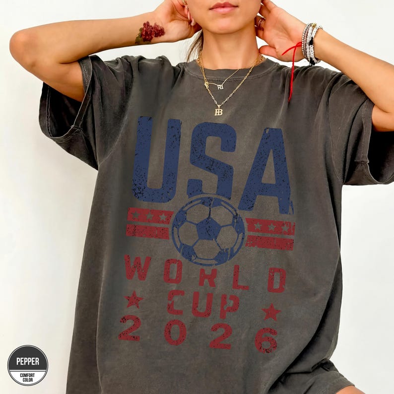 May include: Dark gray t-shirt with the text "USA WORLD CUP 2026" in blue and red. A soccer ball graphic is centered below the word "USA". The shirt has a vintage, distressed look.