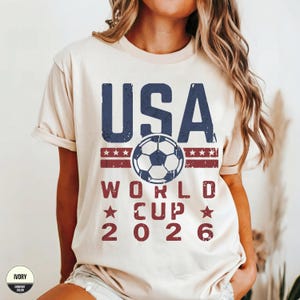 May include: Ivory-colored t-shirt with a distressed graphic design. The design features the word "USA" in navy blue, a soccer ball, and the words "WORLD CUP 2026" in red. The shirt is a comfort color.