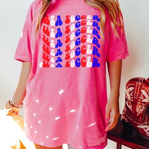 May include: A pink t-shirt with a graphic design of the words "USA SOCCER" repeated in a retro style with red, white, and blue colors. The text is in a wavy font.