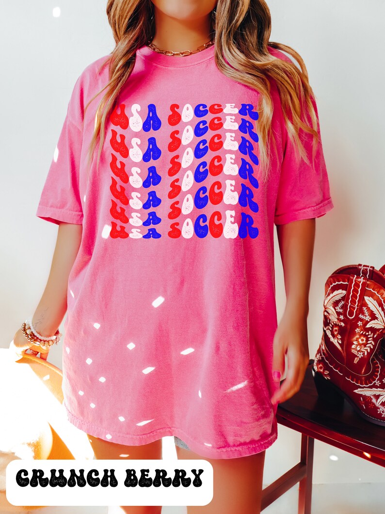 May include: A pink t-shirt with a graphic design of the words "USA SOCCER" repeated in a retro style with red, white, and blue colors. The text is in a wavy font.