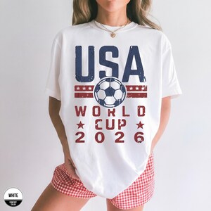 May include: White t-shirt with a USA World Cup 2026 design. The design features the word "USA" in blue, a soccer ball, and the text "WORLD CUP 2026" in red. The shirt is a comfort color.