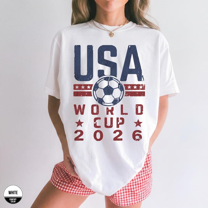 May include: White t-shirt with a USA World Cup 2026 design. The design features the word "USA" in blue, a soccer ball, and the text "WORLD CUP 2026" in red. The shirt is a comfort color.