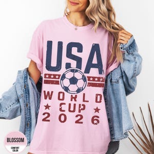 May include: A light pink t-shirt with the text "USA WORLD CUP 2026" in distressed navy blue and red lettering. A soccer ball graphic is centered above the text. The shirt is a comfort color and has rolled-up sleeves.