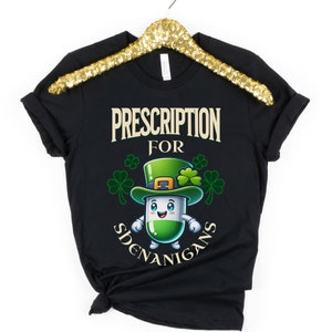 May include: Black t-shirt with a green cartoon pill wearing a leprechaun hat and the text "Prescription for Shenanigans".