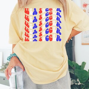 May include: A yellow t-shirt with the words "USA SOCCER" repeated in a retro font in red, white, and blue. The shirt is being worn by a person.