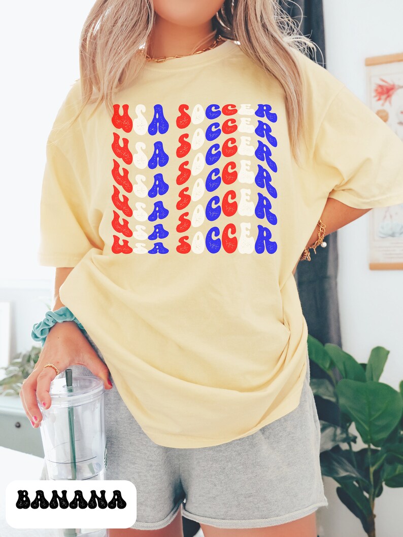 May include: A yellow t-shirt with the words "USA SOCCER" repeated in a retro font in red, white, and blue. The shirt is being worn by a person.