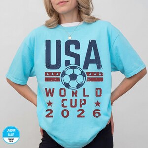 May include: A light blue t-shirt with the text "USA WORLD CUP 2026" in navy blue and red. The design includes a soccer ball and stars, celebrating the upcoming World Cup.