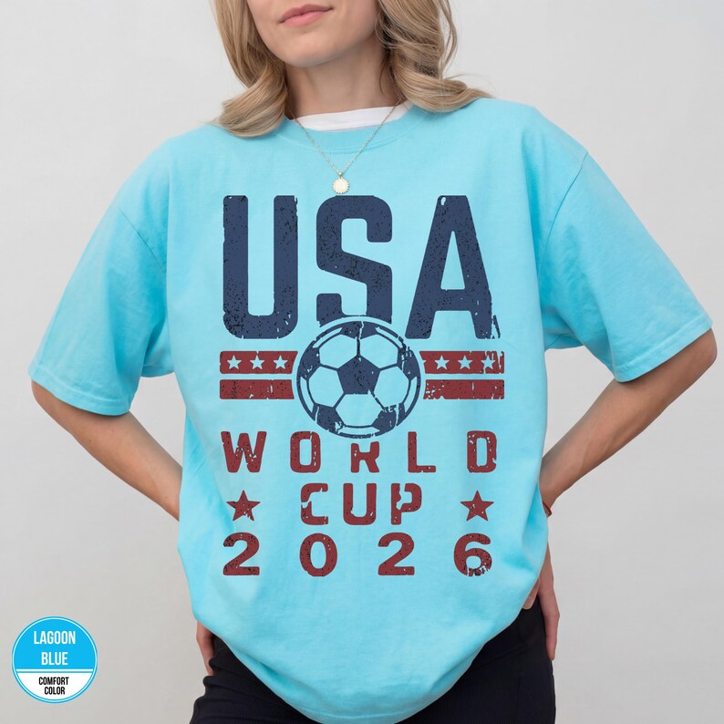 May include: A light blue t-shirt with the text "USA WORLD CUP 2026" in navy blue and red. The design includes a soccer ball and stars, celebrating the upcoming World Cup.