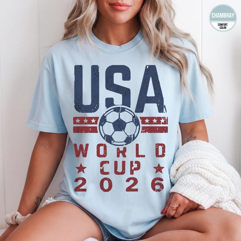 May include: Light blue t-shirt with a distressed graphic. The design features the word "USA" in navy blue above a soccer ball and the text "WORLD CUP 2026" in red. The shirt is made of a soft, comfortable material.