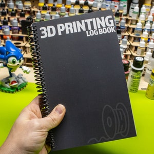 May include: A black spiral bound notebook with the text "3D PRINTING LOGBOOK" on the cover. The notebook has a gray spool of filament icon on the bottom right corner.