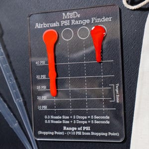 May include: A clear plastic airbrush PSI range finder with a red paint drop on the right side. The range finder has a scale marked in PSI from 10 to 40. The text on the range finder reads "Airbrush PSI Range Finder" and "Range of PSI (Stopping Point) - (+10 PSI from Stopping Point)".