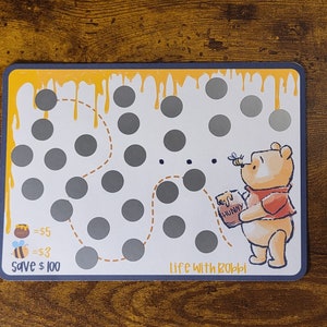 May include: A Winnie the Pooh themed scratch-off card with a yellow and orange border. The card has a picture of Winnie the Pooh holding a jar of honey and a bee flying next to him. The card has a dotted line path with 25 circles that can be scratched off. The card says "Life with Rabbi" at the bottom and "Save $100" at the bottom left corner.
