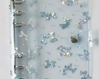May include: A clear plastic binder cover with a silver ring mechanism. The cover has a holographic pattern of silver bows on a white background.