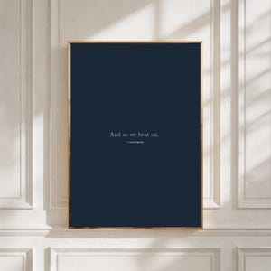 Puede incluir: Impresión artística enmarcada con fondo azul marino y marco dorado. La impresión presenta la cita "And so we beat on." de F. Scott Fitzgerald en texto blanco. La obra de arte se muestra contra una pared blanca.