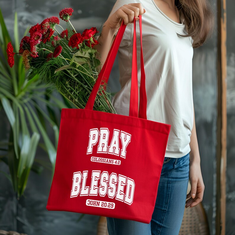 Pode incluir: Uma sacola vermelha com o texto branco "PRAY BLESSED" e refer&ecirc;ncias b&iacute;blicas. A sacola &eacute; segurada por uma pessoa e cont&eacute;m flores vermelhas e vegeta&ccedil;&atilde;o.