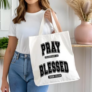 Pode incluir: Uma sacola branca com as palavras "PRAY" e "BLESSED" impressas em letras pretas. O texto "Colossenses 4:2" e "Jo&atilde;o 20:29" tamb&eacute;m est&aacute; impresso na sacola. A sacola &eacute; segurada por uma pessoa vestindo uma camiseta branca e jeans azul.