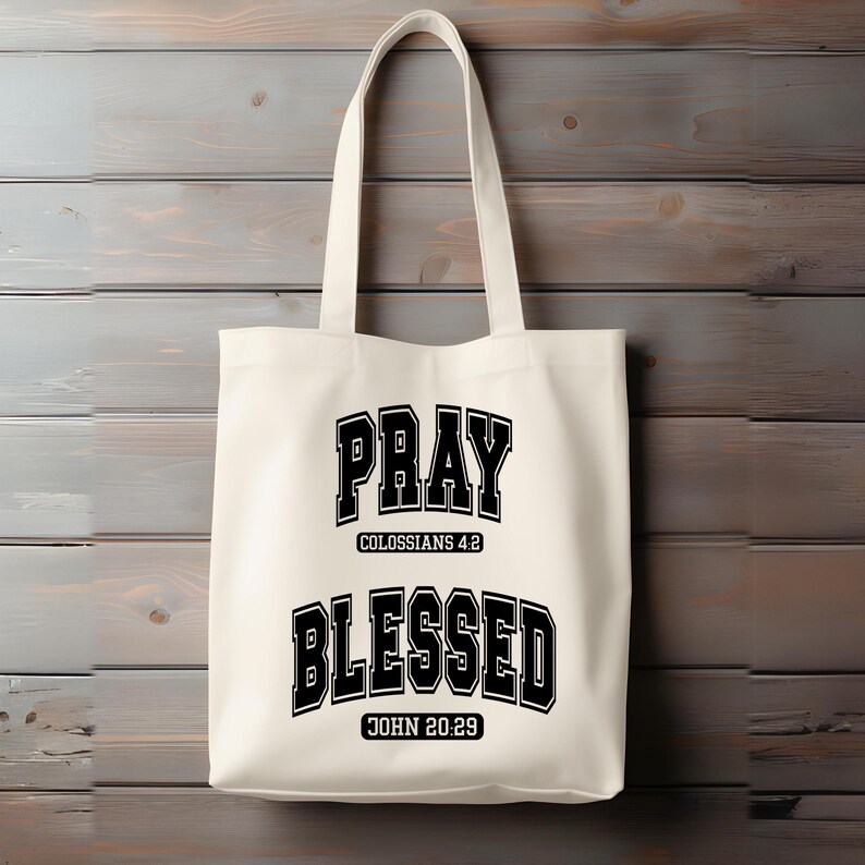Pode incluir: Saco tote creme com texto preto que diz "PRAY" acima de "COLOSSIANS 4:2" e "BLESSED" acima de "JOHN 20:29". O saco tem duas al&ccedil;as e est&aacute; pendurado em um fundo de madeira.