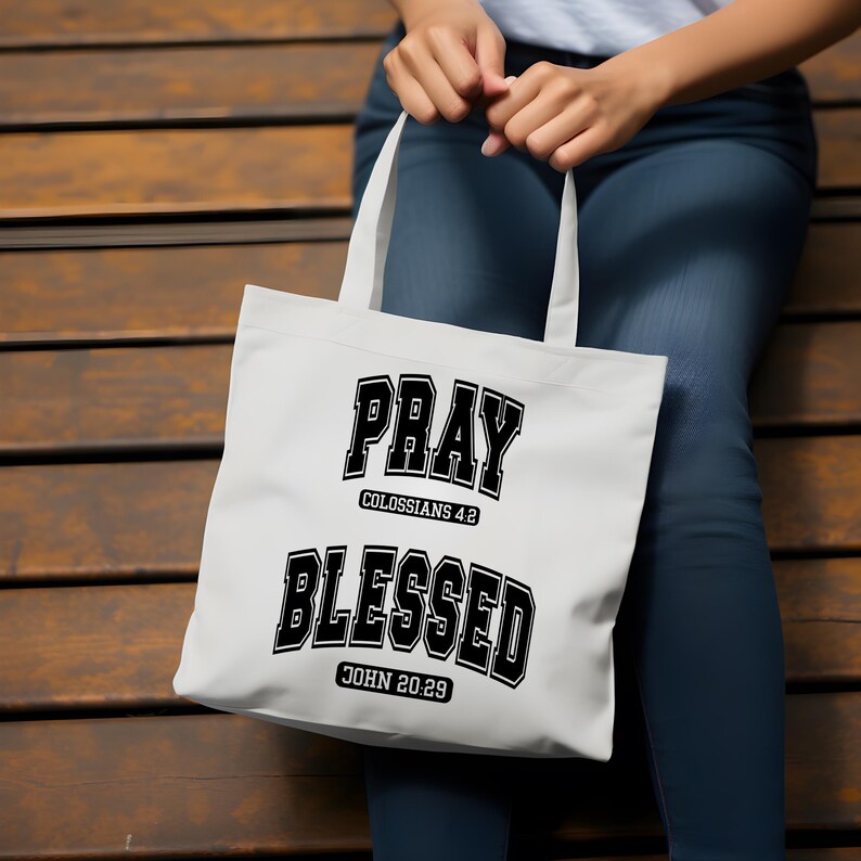 Pode incluir: Bolsa tote branca com texto preto que diz "PRAY" acima de "COLOSSIANS 4:2" e "BLESSED" acima de "JOHN 20:29". A bolsa tem duas al&ccedil;as brancas e &eacute; segurada em frente a um fundo de madeira.