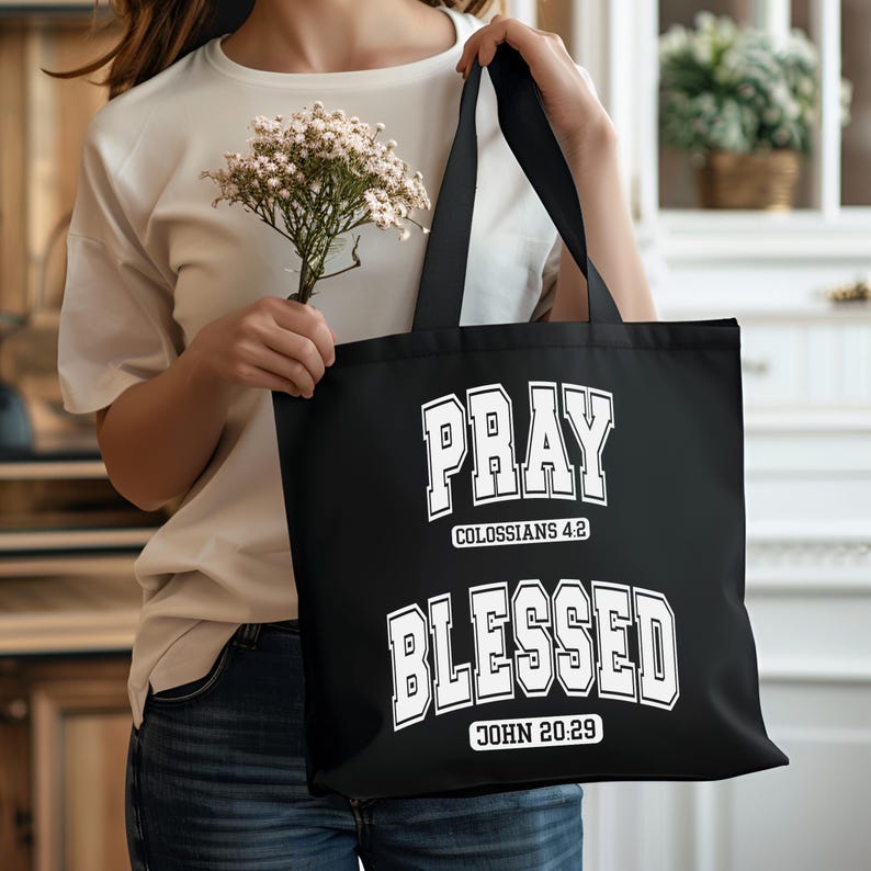 Pode incluir: Bolsa tote preta com as palavras "PRAY" e "BLESSED" em letras brancas. A bolsa tamb&eacute;m inclui os vers&iacute;culos b&iacute;blicos Colossenses 4:2 e Jo&atilde;o 20:29. A bolsa tem duas al&ccedil;as pretas.