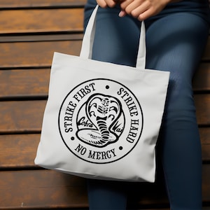 May include: White tote bag with black text and a cobra graphic. The text reads "Strike First Strike Hard No Mercy".