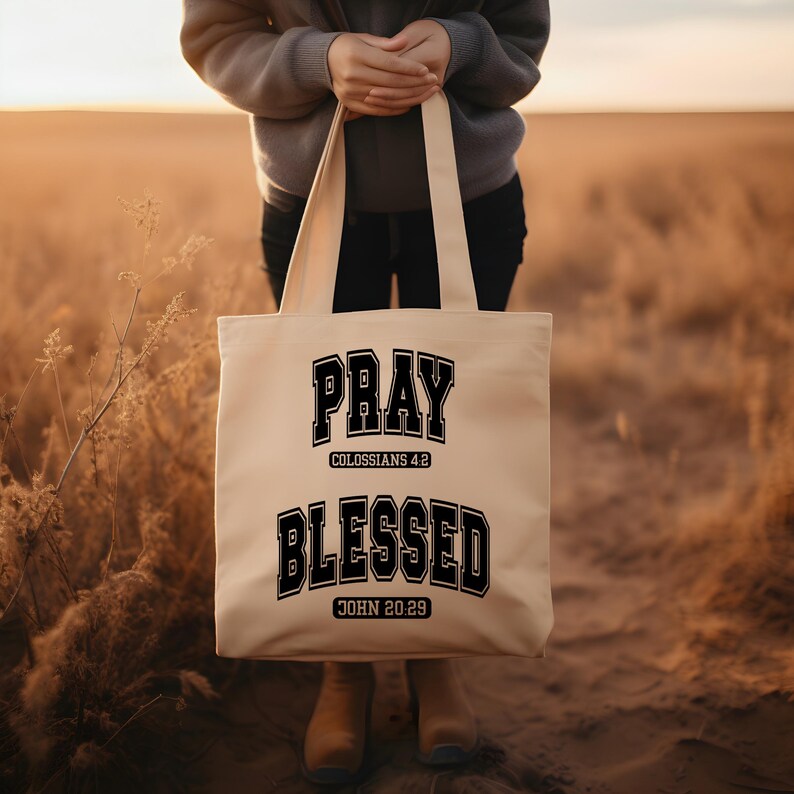 Pode incluir: Uma sacola bege com as palavras "PRAY" e "BLESSED" impressas em preto, com letras em estilo universit&aacute;rio. A sacola tamb&eacute;m apresenta os vers&iacute;culos b&iacute;blicos Colossenses 4:2 e Jo&atilde;o 20:29. A sacola &eacute; segurada por uma pessoa em um campo.