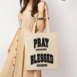 Pode incluir: Uma sacola bege com as palavras "PRAY" e "BLESSED" impressas em preto, em estilo universit&aacute;rio. A sacola tamb&eacute;m apresenta os vers&iacute;culos b&iacute;blicos Colossenses 4:2 e Jo&atilde;o 20:29. A sacola &eacute; segurada por uma pessoa vestindo um vestido bege.