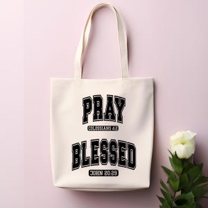 Pode incluir: Bolsa tote creme com as palavras "PRAY" e "BLESSED" em preto, com letras em estilo universit&aacute;rio. A bolsa inclui os vers&iacute;culos b&iacute;blicos Colossenses 4:2 e Jo&atilde;o 20:29. A bolsa tem duas al&ccedil;as e est&aacute; sobre um fundo rosa.