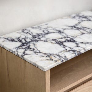 May include: A close-up of a marble countertop with a white base and dark purple veining. The countertop is set on a light-colored wooden cabinet with an open shelf. The marble has a polished finish.